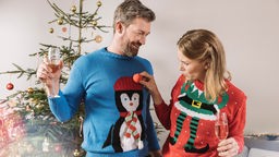 Ein Mann und eine Frau stehen mit Weihnachtspullis vor einem Weihnachtsbaum.