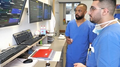 Zwei Männer in blauer Krankenhauskleidung schauen auf Monitore, die an der Wand hängen