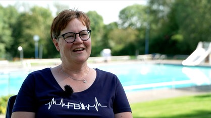 Eine Frau mit kurzen roten Haaren und Brille steht vor einem Schwimmbecken