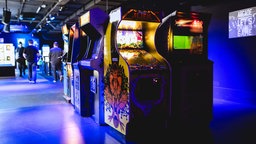 Mehrere Arcade-Spieleautomaten stehen nebeneinander aufgereiht in einer Halle. 