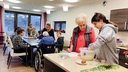 Im Vordergrund kneten eine Jugendliche und eine Seniorin Keksteig, im Hintergrund sitzt eine Gruppe Senioren an einem langen Tisch