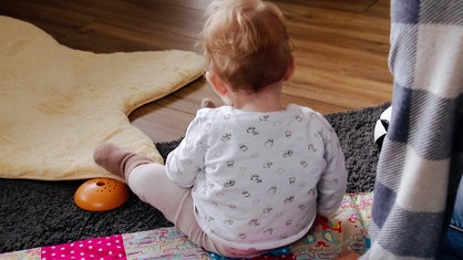 Ein kleines Kind sitzt auf einer Decke auf dem Boden, es ist nur von hinten zu sehen