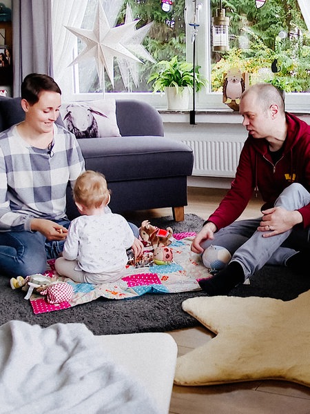 Eine Frau und ein Mann spielen am Boden mit einem Kleinkind, das Kind ist nur von hinten zu sehen