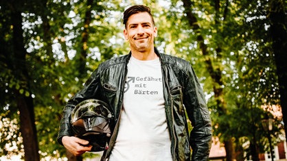 Ein Mann mit schwarzer Lederjacke. Darunter trägt er ein weißes T-Shirt mit der Aufschrift "Gefährten mit Bärten" und einen Motorradhelm unter dem rechten Arm