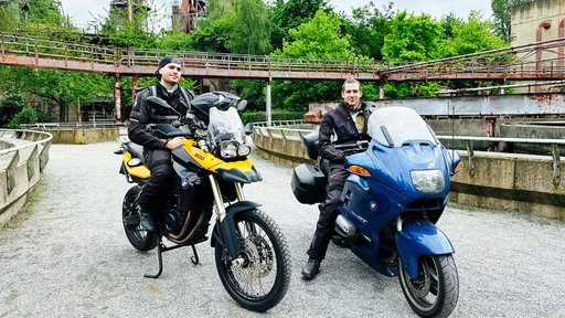 Zwei Männer in Motorradkluft sitzen auf einem jeweils blauen und gelben Motorrad und lächeln in die Kamera