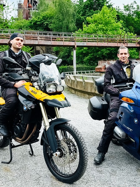 Zwei Männer in Motorradkluft sitzen auf einem jeweils blauen und gelben Motorrad und lächeln in die Kamera