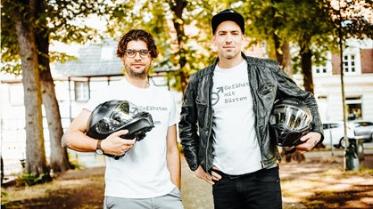 Zwei Männer mit weißen T-Shirts mit der Aufschrift "Gefährten mit Bärten", die beide einen Motorradhelm unter dem Arm tragen