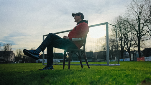 Björn Freiburg sitzt auf einem Holzstuhl auf einem Fußballfeld mit dem Tor und der untergehenden Sonne im Hintergrund.