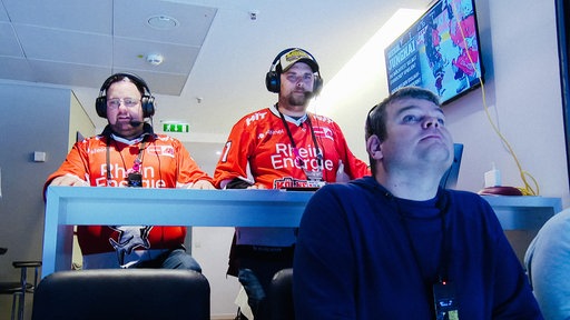 Blindenreporter Björn Naß und sein Kollege stehen mit Headset an einem Pult den Blick auf ein Eishockeyfeld hinter der Kamera gerichtet. Vor ihnen sitzt ein blinder Eishockey-Fan und hört ihrer Reportage zu