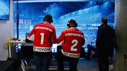 Zwei Männer in Trikots mit der Aufschrift "Blindenreportage", in einer Arena.