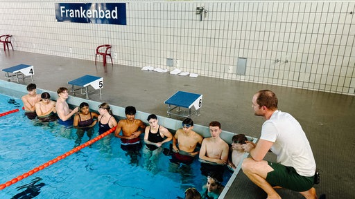 Ein mittelalter Mann mit braunen Haaren am Rande eines Schwimmbeckens. Im Wasser einige junge Leute in Schwimmklamotten