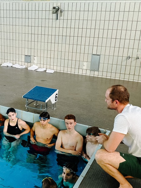 Ein mittelalter Mann mit braunen Haaren am Rande eines Schwimmbeckens. Im Wasser einige junge Leute in Schwimmklamotten