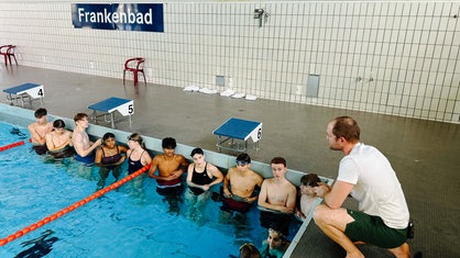 Ein mittelalter Mann mit braunen Haaren am Rande eines Schwimmbeckens. Im Wasser einige junge Leute in Schwimmklamotten