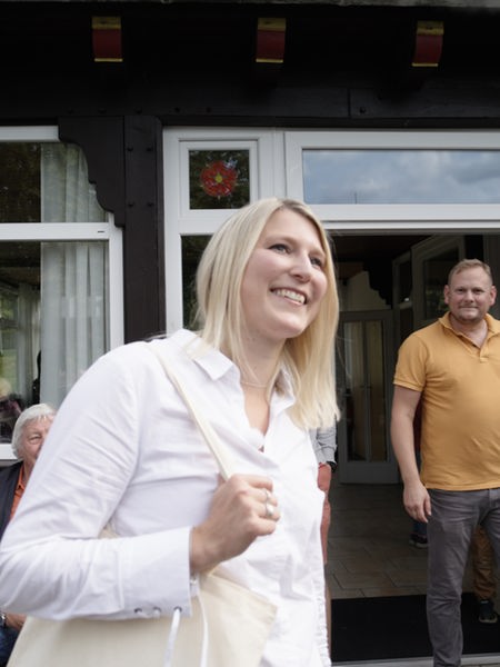 Laura Schuster ist eine Frau mit blonden, schulterlangen Haaren und einer weißen Bluse. Sie steht in der Mitte des Bildes und lächelt. Hinter ihr sind ein Fachwerkhaus und vier Personen zu sehen.