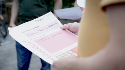 Eine Person hält einen weiß-pinken Zettel in der Hand auf dem "Name für Projekt" steht und mehrere Textfelder zu sehen sind.