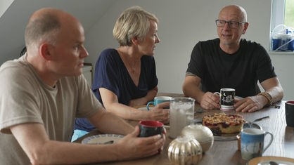 Die drei Freiwilligen sitzen am Kaffeetisch mit Kaffee und Kuchen und unterhalten sich.