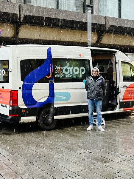 Ein weißer Kastenwagen in einer Einkaufsstraße. Davor ein Mann in Regenjacke. Links ein blauer Anhänger