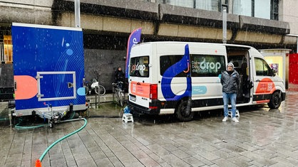 Ein weißer Kastenwagen in einer Einkaufsstraße. Davor ein Mann in Regenjacke. Links ein blauer Anhänger