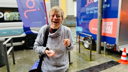 Eine Frau lächelt in die Kamera. Sie hat blonde Haare. Im Hintergrund ein Kastenwagen und ein Anhänger mit blauer Farbe