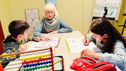 Eine Frau mit grauen Haaren und Brille sitzt mit einem Jungen und einem Mädchen im Grundschulalter an einem Tisch, sie beugen sich über Arbeitshefte