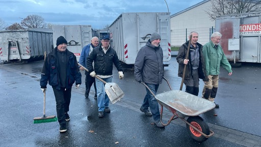 Sechs Männern laufen über einen Recyclinghof, einer schiebt Schubkarre, andere tragen Schaufel oder Besen