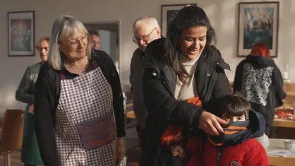 Angela Müllers steht neben einer Frau und beobachtet sie lächelnd dabei, wie sie ihrem Sohn fröhlich den Schal auszieht.