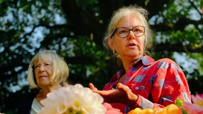 Eine blonde Frau mit Bille in einem rot-blau karierten Hemd steht in einem Garten