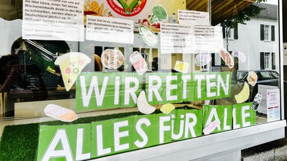 Ein Schaufenster, in dem mehrere Plakate und Schilder hängen. Auf einem steht "Wir retten alles für alle."