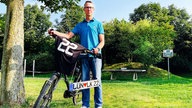 Ein Mann im blauen Poloshirt und Brille hält ein Fahrrad. Auf dem Lenker liegt ein Trikot mit der Nummer 22. Darunter ein Kennzeichen "LÜN Herz LK 22" 