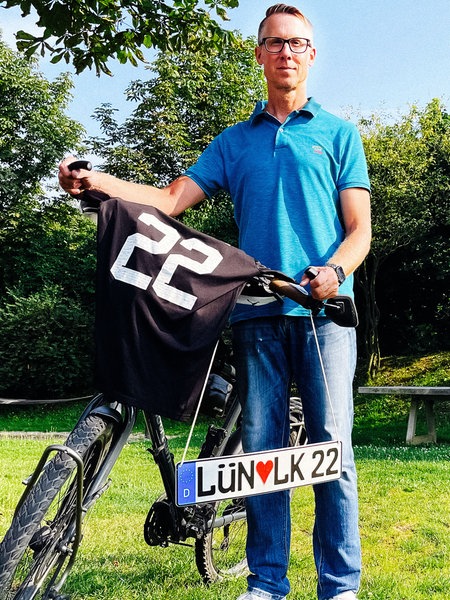 Ein Mann im blauen Poloshirt und Brille hält ein Fahrrad. Auf dem Lenker liegt ein Trikot mit der Nummer 22. Darunter ein Kennzeichen "LÜN Herz LK 22" 