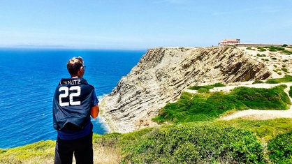 Ein Mann mit Brille blickt aufs Meer, rechts daneben sind Felsklippen und ein Haus. Der Mann hat ein Trikot mit der Aufschrift L. Kautz und der Nummer 22 auf dem Rücken.