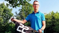 Ein Mann im blauen Poloshirt und Brille hält ein Fahrrad. Auf dem Lenker liegt ein Trikot mit der Nummer 22. Darunter ein Kennzeichen "LÜN Herz LK 22" 