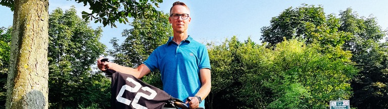 Ein Mann im blauen Poloshirt und Brille hält ein Fahrrad. Auf dem Lenker liegt ein Trikot mit der Nummer 22. Darunter ein Kennzeichen "LÜN Herz LK 22" 