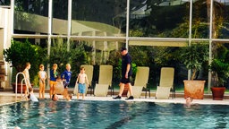 Ein Mann und mehrere Kinder stehen an einem Schwimmbeckenrand. 