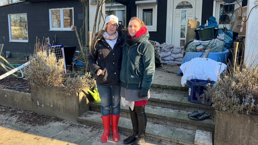 Zwei Frauen in Winterjacken stehen vor einem Haus. Sandsäcke und Werkzeuge liegen vor dem Eingang.