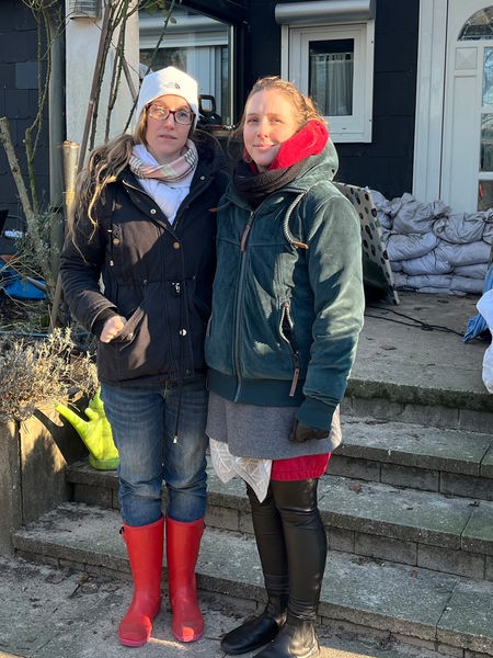 Zwei Frauen in Winterjacken stehen vor einem Haus. Sandsäcke und Werkzeuge liegen vor dem Eingang.
