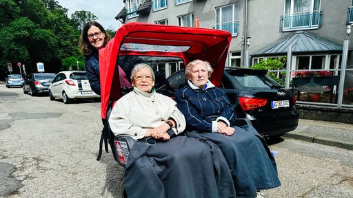 Eine Rikscha, auf der zwei Seniorinnen sitzen. Hinten lächelt eine Frau, die das Fahrrad fährt.