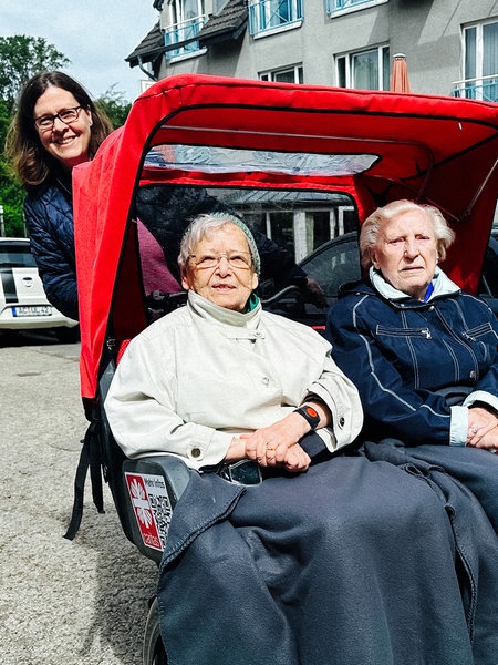 Eine Rikscha, auf der zwei Seniorinnen sitzen. Hinten lächelt eine Frau, die das Fahrrad fährt.