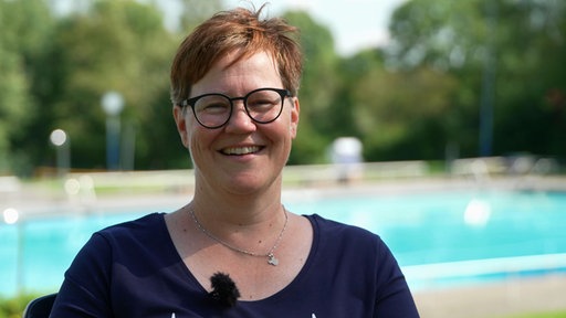 Eine Frau mit kurzen roten Haaren und Brille steht vor einem Schwimmbecken