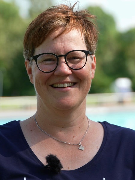 Eine Frau mit kurzen roten Haaren und Brille steht vor einem Schwimmbecken