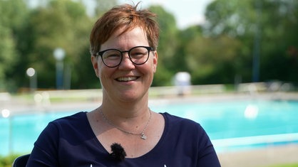 Eine Frau mit kurzen roten Haaren und Brille steht vor einem Schwimmbecken