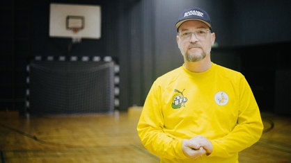 Ein Mann mit grauem Bart, Brille und, dunkelblauer Kappe schaut in Richtung Kamera. Er trägt ein gelbes Trikot. Im Hintergrund ein Fußballtor