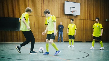 Vier jugendlich-aussehende Jungen in einer Sporthalle. Sie tragen gelbe Trikots. Es findet ein Zweikampf um einen Ball statt