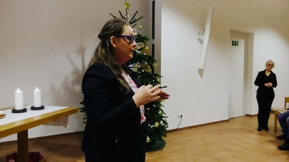 Eine Frau steht vor einem Weihnachtsbaum. Am rechten Bildrand sieht man mehrere Personen sitzen und eine Frau die steht