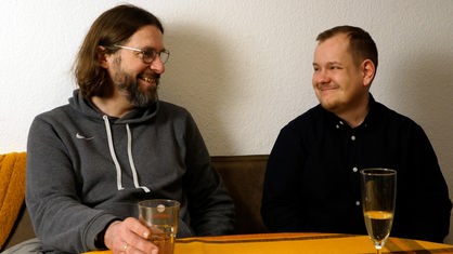 Zwei Männer lächeln sich an, sie sitzen einer Bank an einem Tisch.