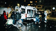 Ein weißer Lieferwagen steht nachts im Regen an einer Straße. Daneben stehen mehrere Menschen, die der Kamera den Rücken zuwenden