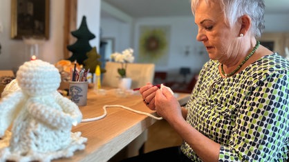 Eine Frau mit grauen Haaren sitzt an einem Tisch und hält eine Häkelnadel sowie weiße Wolle in der Hand