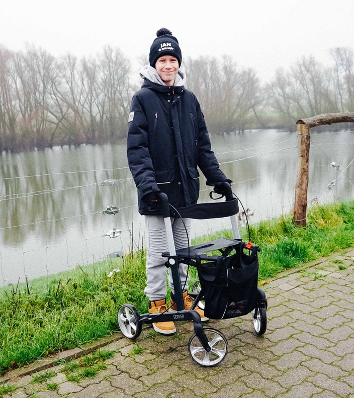 Ein Junge steht mit einem Rolator am Rande eines Gehwegs. Im Hintergrund ist ein Fluss und mehrere Bäume. 