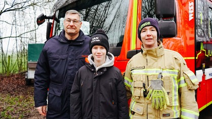 Ralf Benkel, Jan Luca und Nadine Hollenders stehen zu dritt Nebeneinander in einem Waldstück vor einem Feuerwehrauto. 