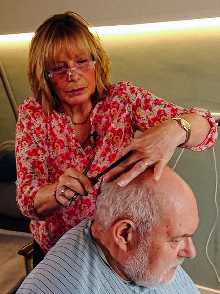 Im Vordergrund sitzt Hospizpatient Jürgen mit einer Friseurschürze auf einem Rollstuhl. Dahinter steht Marion Hebel und schneidet ihm die Haare. Im Hintergrund ist das Hospizbett zu sehen.